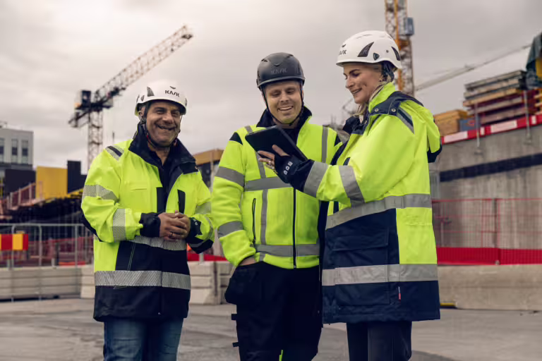 Tre personer på en byggeplass som arbeider med KMA (HMS/KS). De har på seg verneutstyr som hjelm og refleksvester, og de ser ut til å diskutere sikkerhet og kvalitet på arbeidsplassen.
