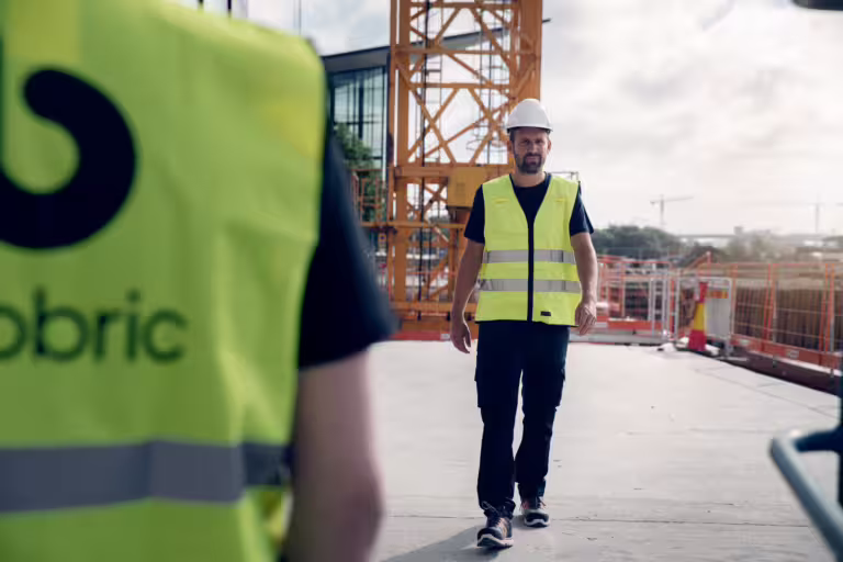 Mann med gul refleksvest og hjelm som går på en byggeplass.