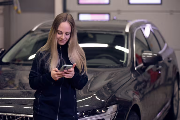 En kvinna står bredvid sin bil och kollar milersättning i mobilen.